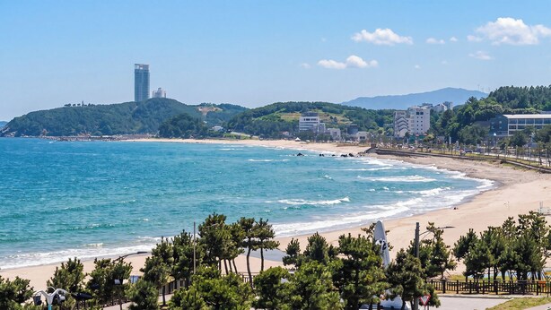 Una playa costera con una orilla arenosa, mar azul cristalino y edificios y colinas al fondo.