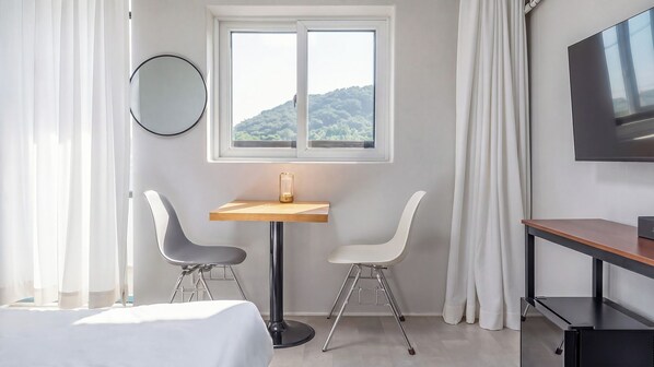 Quarto de hotel com uma cama, uma mesa com duas cadeiras, uma televisão, uma janela com vista para áreas verdes e um espelho redondo na parede.