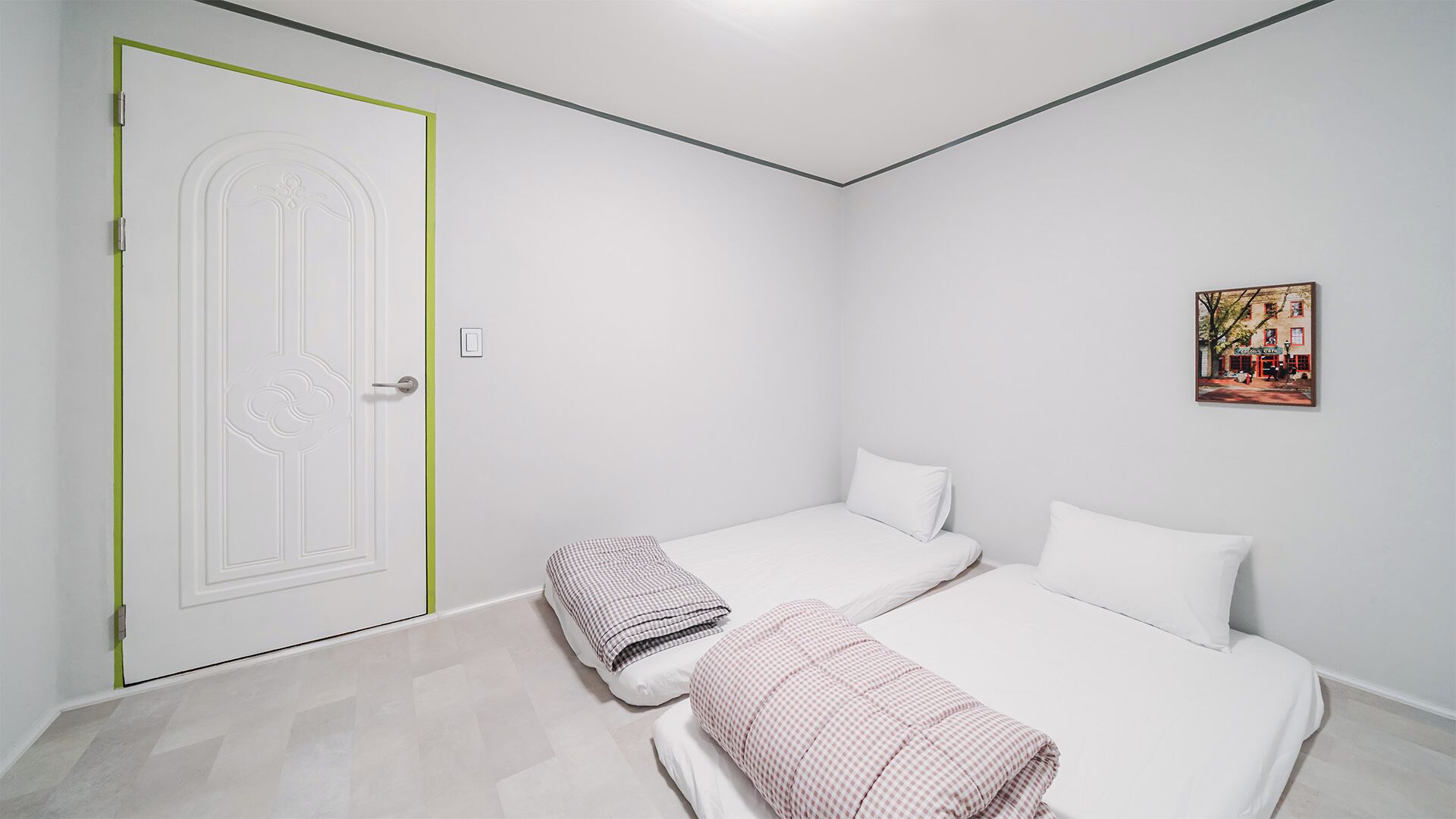 A minimalist room with two beds, a door with a green accent, and a small wall-mounted picture.
