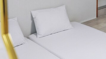 Una cama individual con ropa blanca y almohadas en una habitación con pared lisa y suelo de madera.