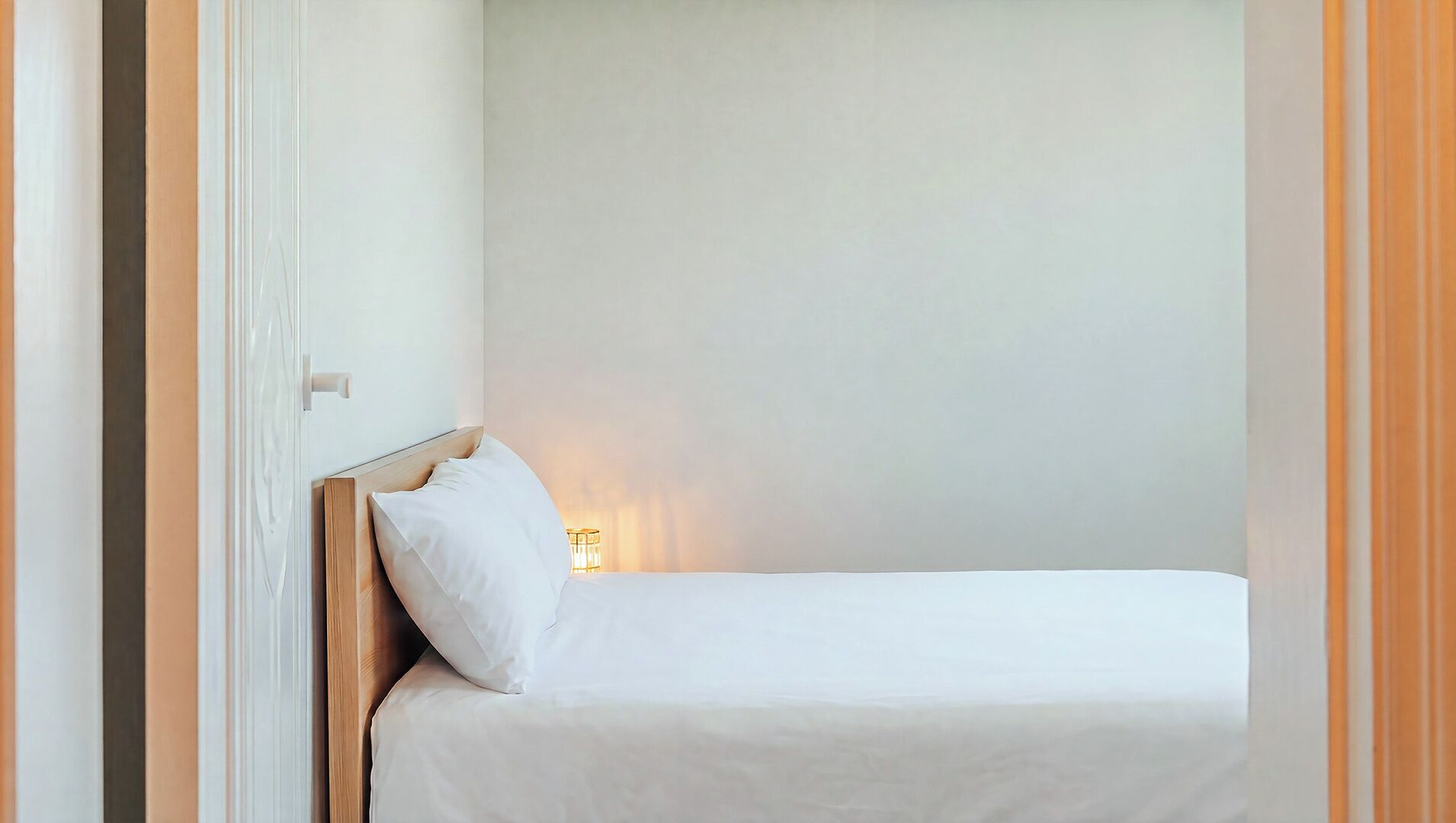 Een eenpersoonsbed met een houten hoofdbord, een wit kussen en een bedlampje in een kamer met effen muren.