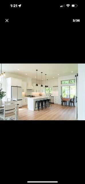 A modern kitchen with white cabinetry, a central island, pendant lighting, and a dining area with a wooden table and chairs. - Sunrise Sunset Bayview Home (Homer)