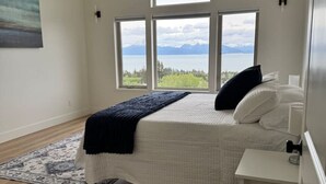 Quarto com cama, mesa de cabeceira, janela com vista para montanhas e água, e um quadro na parede.