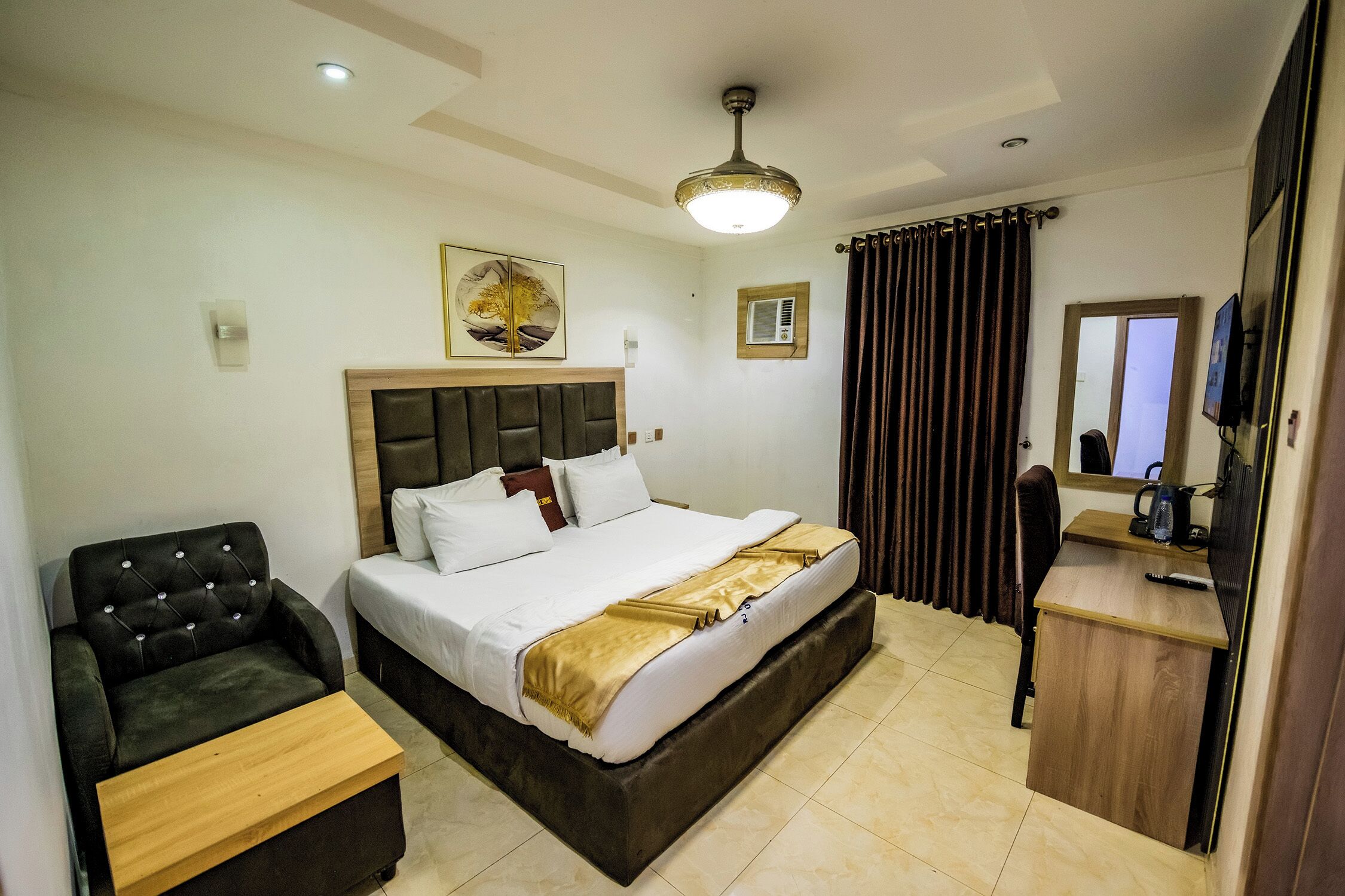 Une chambre d’hôtel avec un lit, un bureau, une chaise, un ventilateur de plafond, un tableau au mur et une fenêtre avec des rideaux.