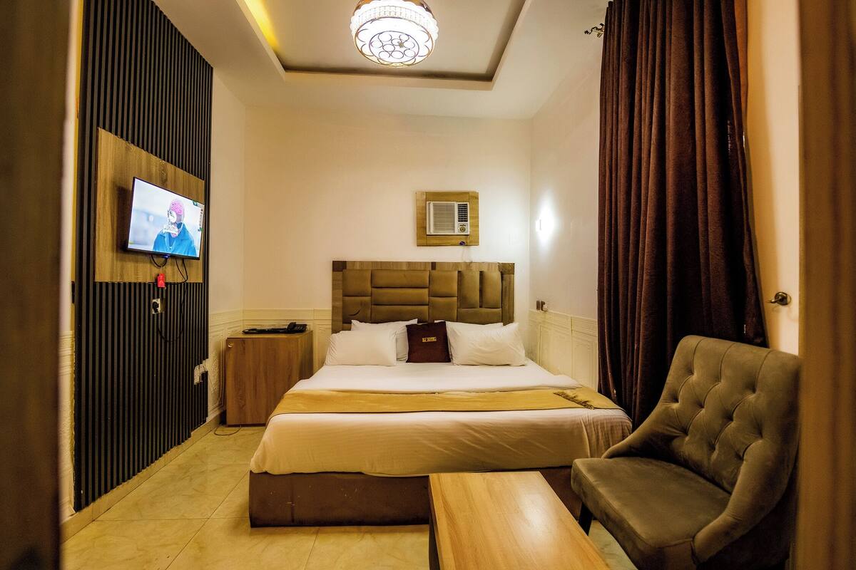 Camera d'albergo con un letto, una televisione, un comodino, una sedia e una lampada a sospensione.