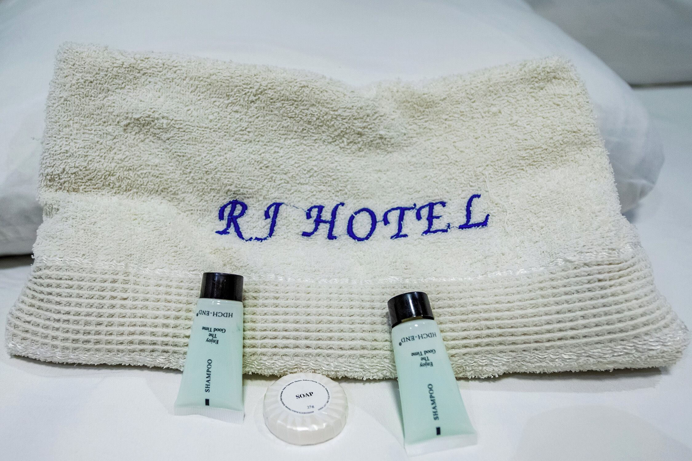 A white towel with 'RJ HOTEL' embroidered in blue, two bottles of shampoo and conditioner, and a soap bar.