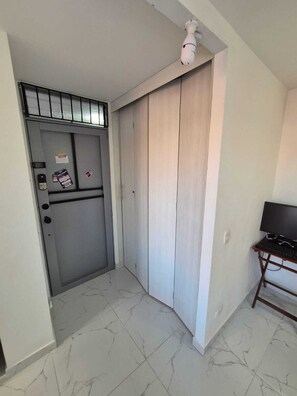Ein Zimmer mit einer grauen Tür, einem kleinen Schreibtisch mit einem Computer und einem Fliesenboden.