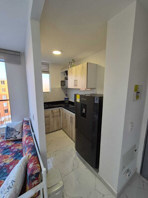 Eine kleine Küche mit Kühlschrank, Mikrowelle und Schränken, eine gemütliche Sitzecke mit einem bunten Kissen und ein Fenster mit Blick auf Gebäude.
