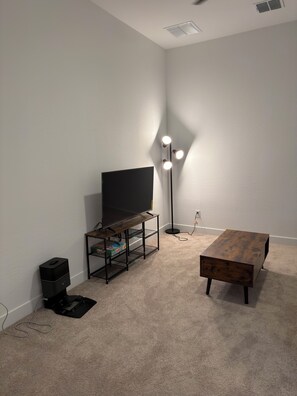 Ein Zimmer mit einem Flachbildfernseher auf einem Holzbodenständer, einer Stehlampe mit drei Leuchten, einem kleinen schwarzen Lautsprecher und einem Holztisch.