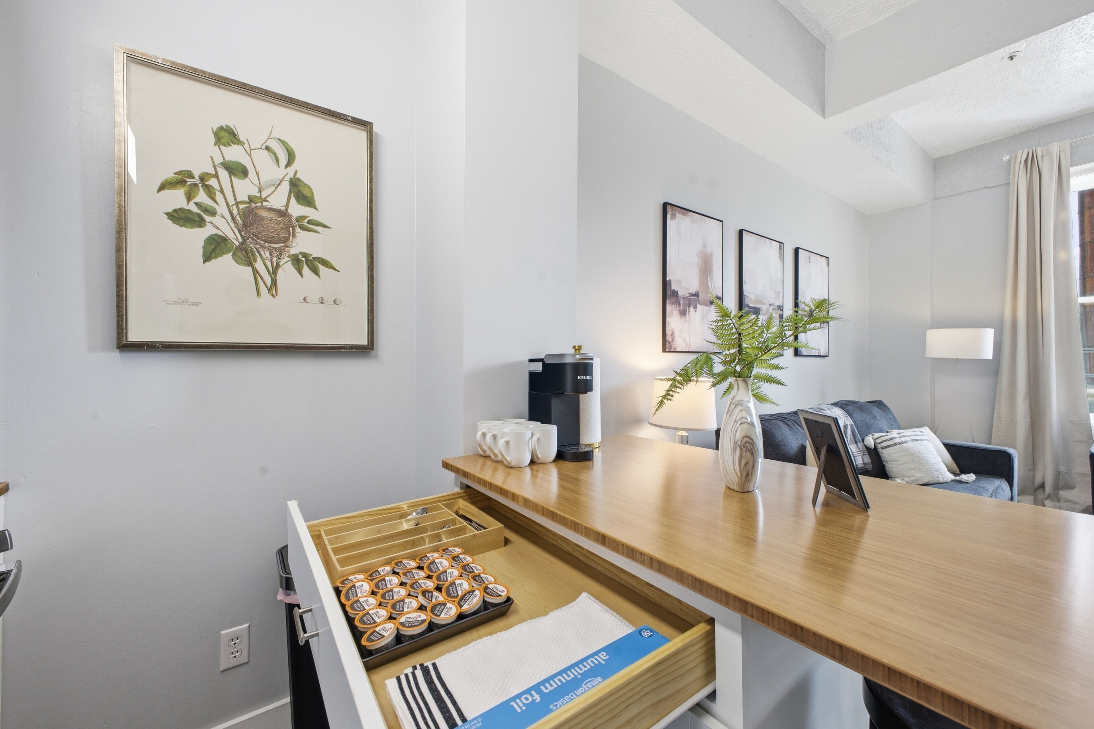 Een moderne hotelkamer met een houten bureau, een koffiezetapparaat, een plant en een ingelijste botanische illustratie aan de muur.