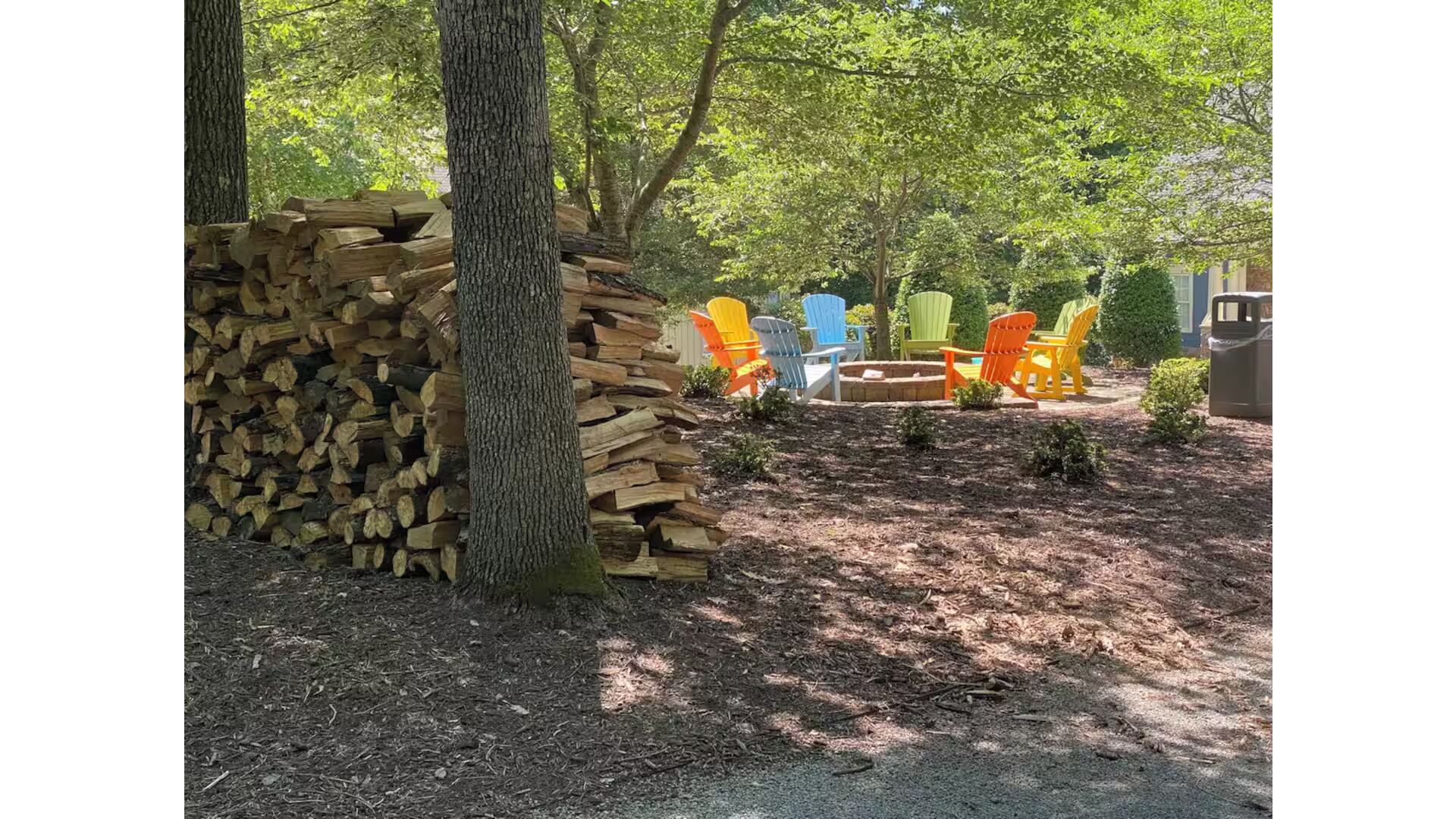 A stack of firewood, outdoor chairs, and a tree in a wooded area.