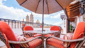 Eine Terrasse mit Korbstühlen, einem Tisch und einem Sonnenschirm. Im Hintergrund ein Glockenturm und ein Gebäude mit einer Kuppel.