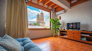 Ein Wohnzimmer mit einer Couch, einem Fernseher, einem Holzschrank und einem Fenster mit Blick auf ein historisches Gebäude.