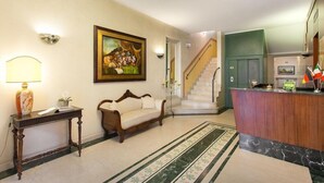 A hotel lobby with a reception desk, a sofa, a painting, a staircase, and a chandelier. - Hotel Gambara (Gambara)