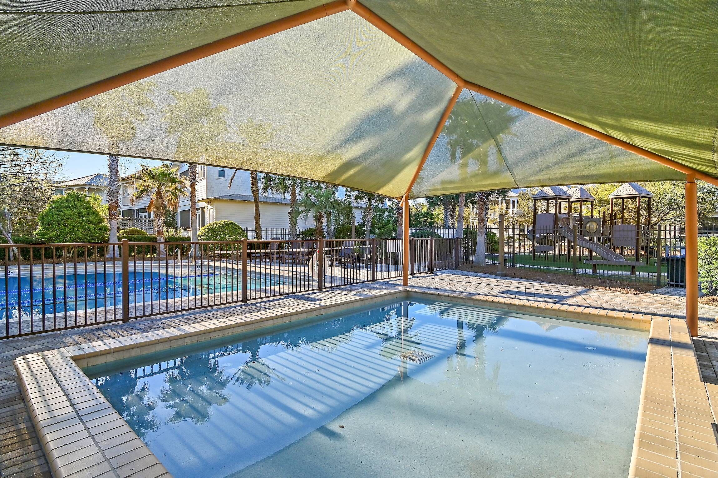 A swimming pool with a shaded area and a fenced playground in the background.