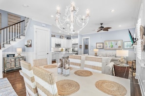 A spacious living area with a chandelier, wicker furniture, and a kitchen in the background. - Beach Blessings | 4BR 30A | Sleeps 8 | 6 Seater Golf Cart + Bikes (Santa Rosa Beach)
