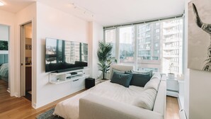 Ett modernt sovrum med en stor säng, en platt-TV som är monterad på väggen, en krukväxt och utsikt över en stadssiluett genom stora fönster.