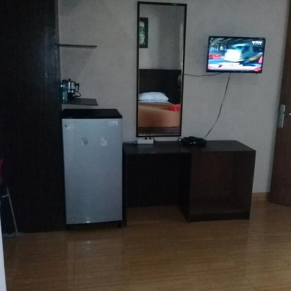 A compact room with a small refrigerator, a desk, a mirror, and a television mounted on the wall. - RF Hotel (Cisarua)