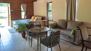 Um quarto aconchegante com teto de madeira, uma cama com almofadas amarelas, um sofá com almofadas verdes, uma mesa redonda e plantas em vasos.