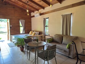 A cozy room with a wooden ceiling, a bed with yellow pillows, a sofa with green cushions, a round table, and potted plants. - Rain Forest Cabin with Jacuzzi (La Fortuna)