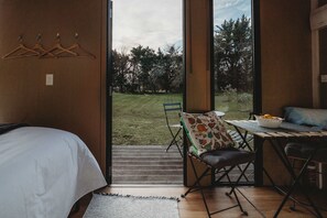 A bedroom with a bed, a wooden table with a bowl of fruit, a chair, and a view of a grassy yard through a large door.