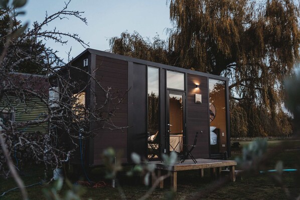 A small cabin with a deck, a visible interior with a bed and a chair, and a large tree in the background.