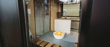 Cocina compacta con puerta de vidrio, un lavabo blanco y una mesa de madera con cesta de frutas.