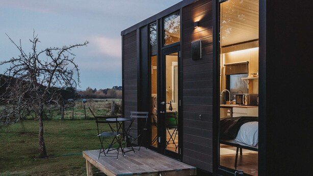 A small, dark wooden cabin with a wooden deck, a table, and chairs outside. Inside, there is a bed and a kitchenette.