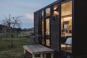 A small, dark wooden cabin with a wooden deck, a table, and chairs outside. Inside, there is a bed and a kitchenette.
