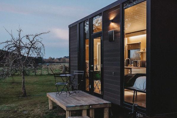 A small, dark wooden cabin with a wooden deck, a table, and chairs outside. Inside, there is a bed and a kitchenette.