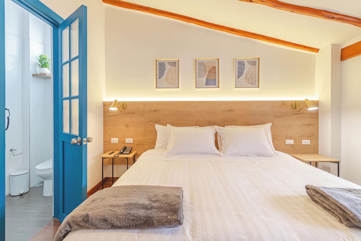 Quarto com cabeceira de madeira, roupa de cama branca, mesa de cabeceira com abajur e uma obra de arte na parede.