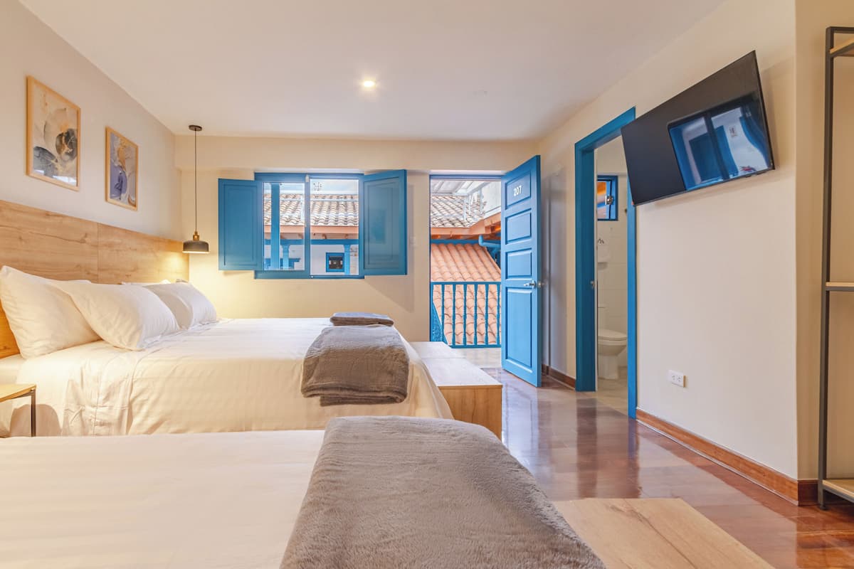 Quarto com duas camas, cabeceira de madeira, um banco, televisão e banheiro visível por uma porta aberta.