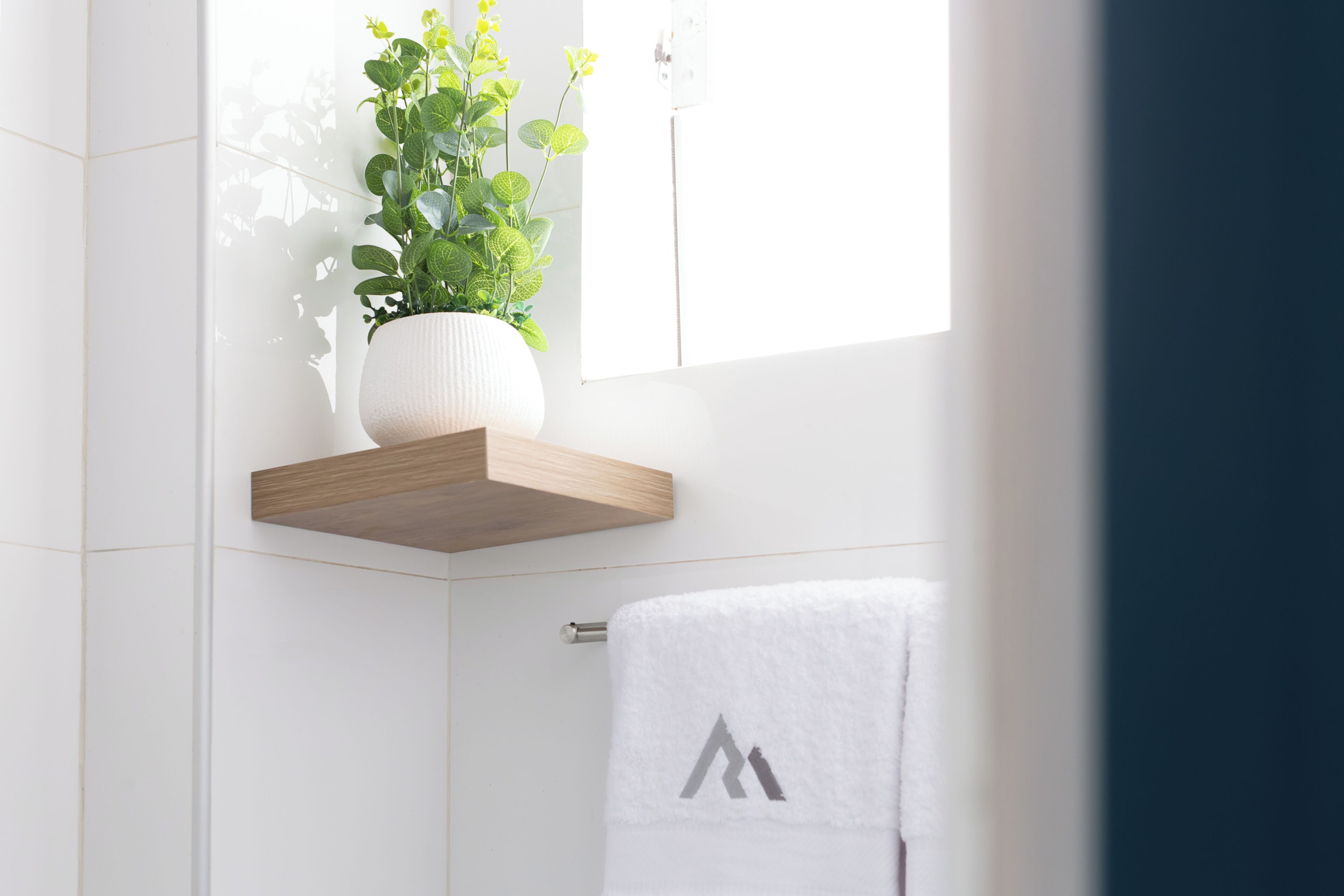 A bathroom with white tiles, a wooden shelf holding a potted plant, and a towel with a logo.