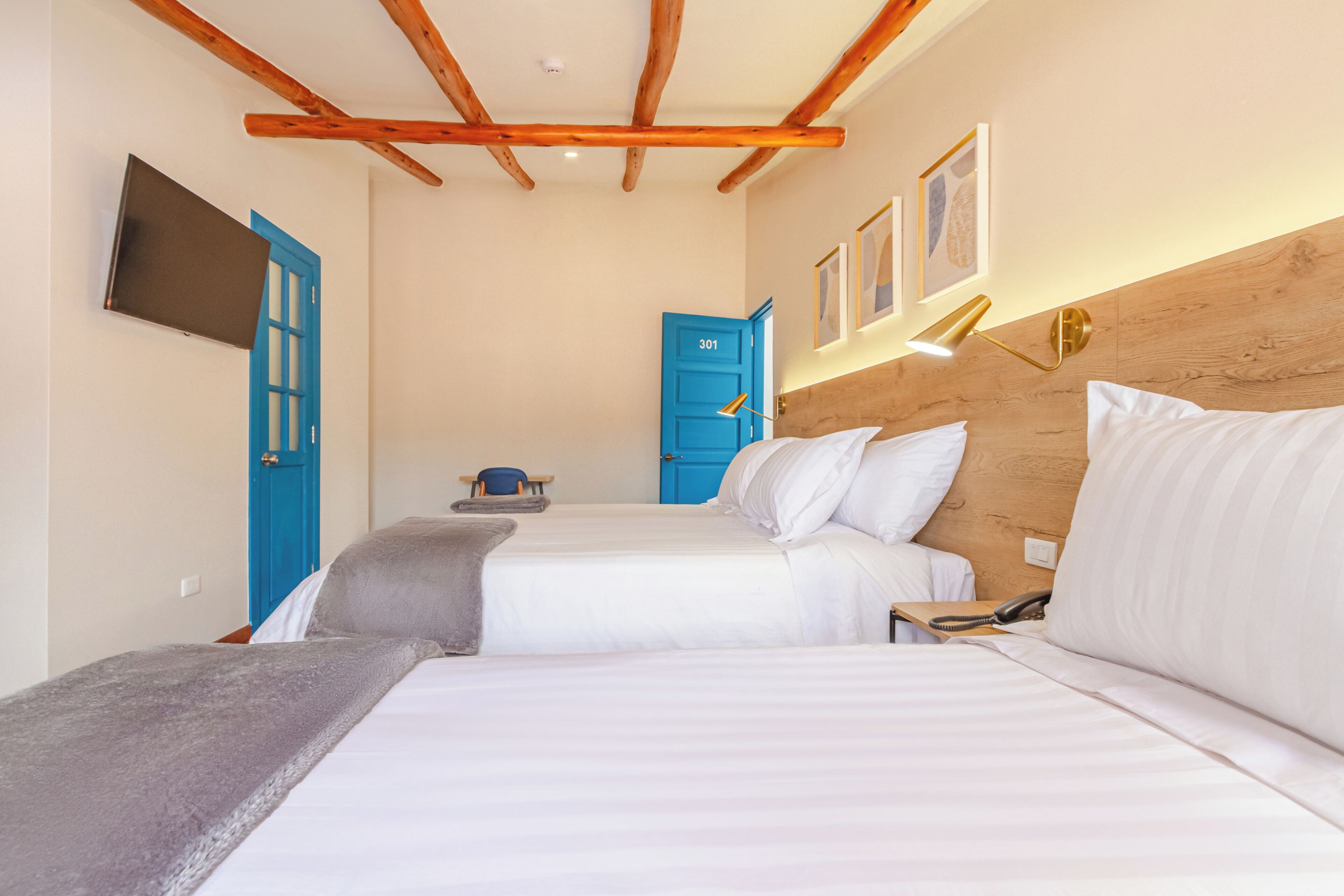 Quarto com duas camas, televisão na parede, porta azul e teto de madeira.