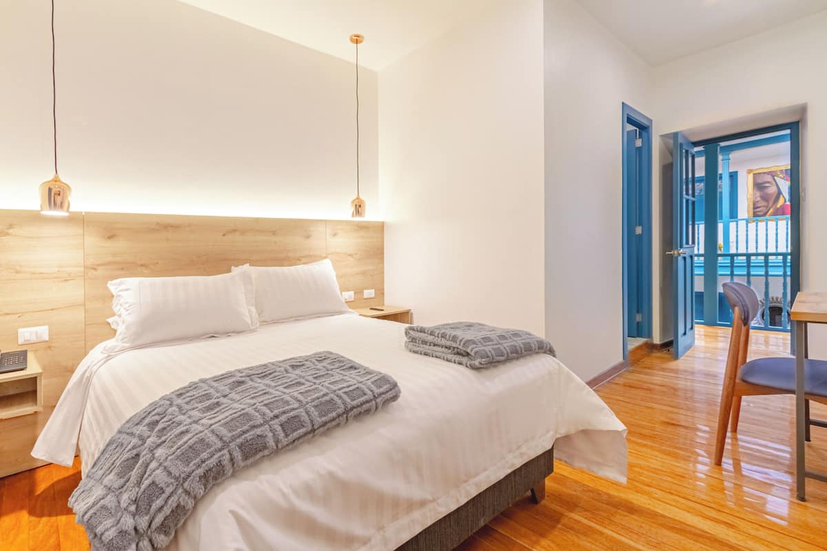 Quarto com cabeceira de madeira, cama com roupa de cama branca e um cobertor cinza, sobre piso de madeira.