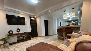 A living room with a brown leather sofa, a flat-screen TV mounted on the wall, a dining area with a table and chairs, and a kitchen area with pendant lights. - Calla House Arenal 3BR (La Fortuna)