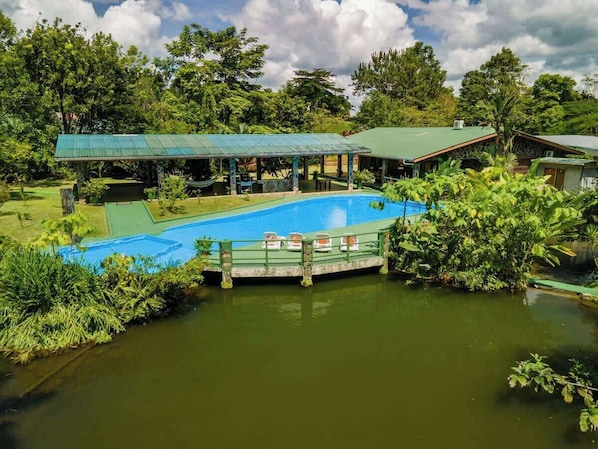 A pool with a wooden deck, surrounded by lush greenery and a covered pavilion.