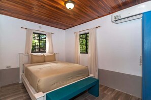 Ein Schlafzimmer mit einem Bett, einer Bank, zwei mit Vorhängen versehenen Fenstern, einer Holzbalkendecke und einer Klimaanlage.