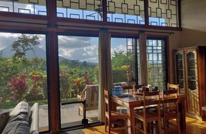 A dining area with a table and chairs, a sofa, and a view of a mountainous landscape through large windows. - Volcano View Oasis Arenal 2BR (San Ramón)