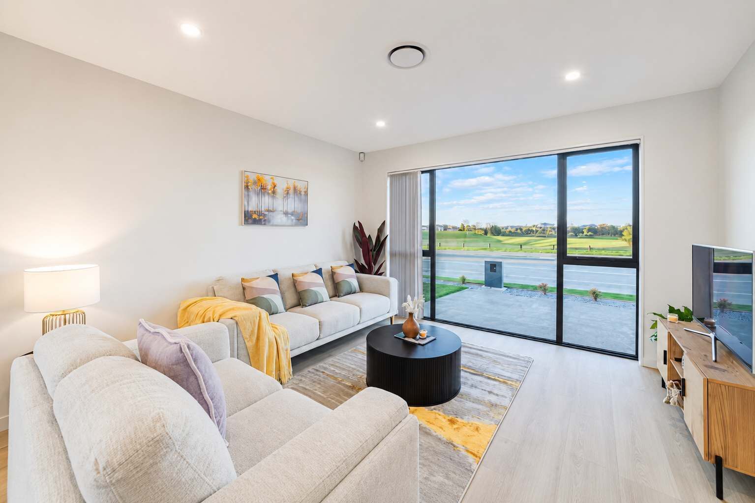 A modern living room with a sofa, a coffee table, a TV, and a large window offering a view of a road and greenery.
