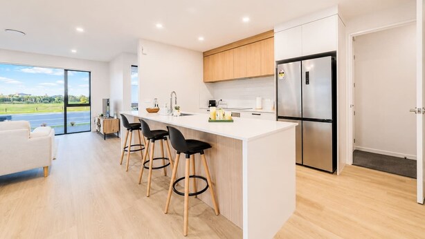 Una cocina moderna con una isla grande, taburetes de bar, un refrigerador de acero inoxidable y vistas a un paisaje verde a través de un ventanal.
