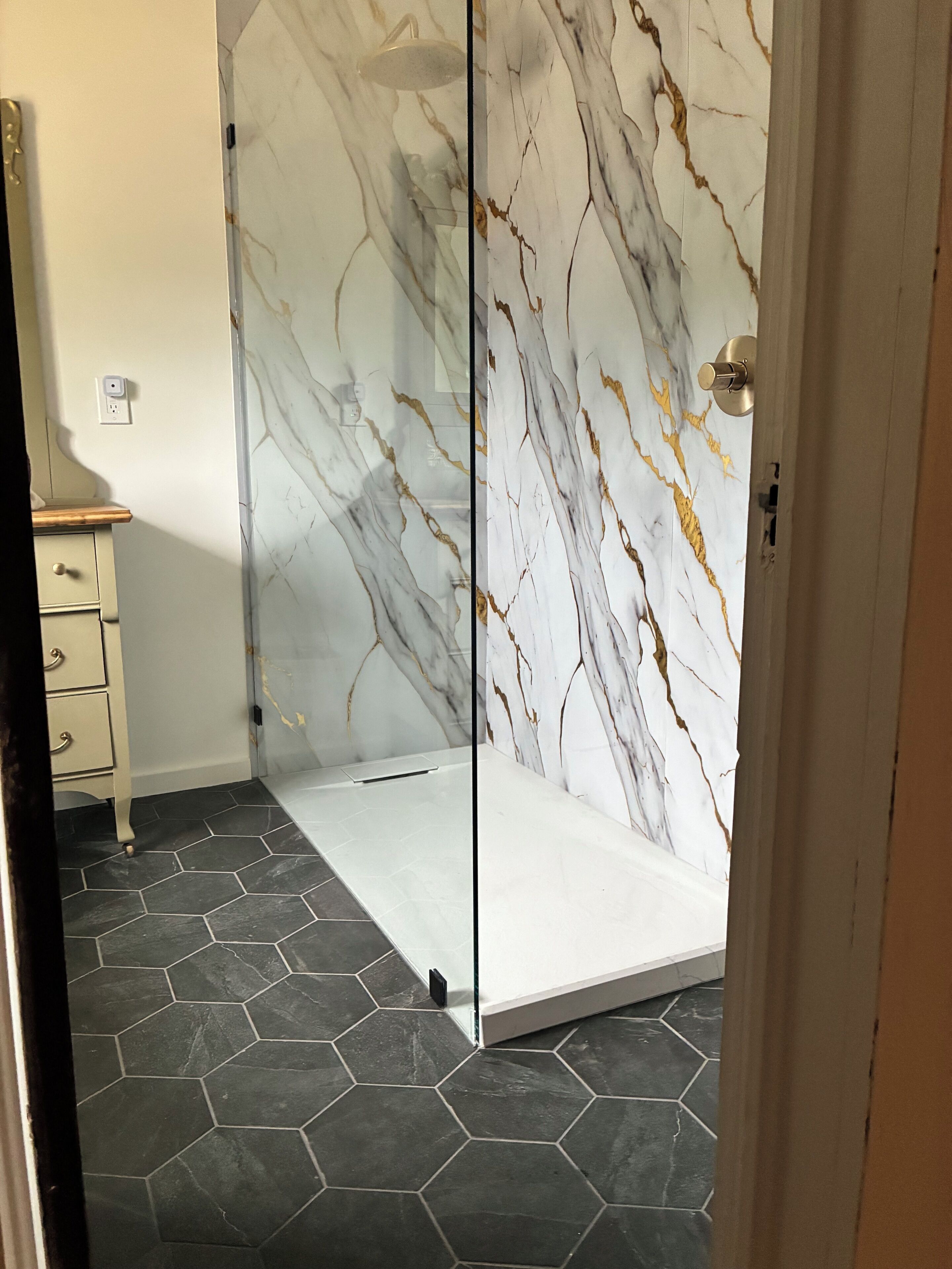 A bathroom with a glass partition, hexagonal dark tiles, a white marble wall with gold veins, and a beige cabinet.