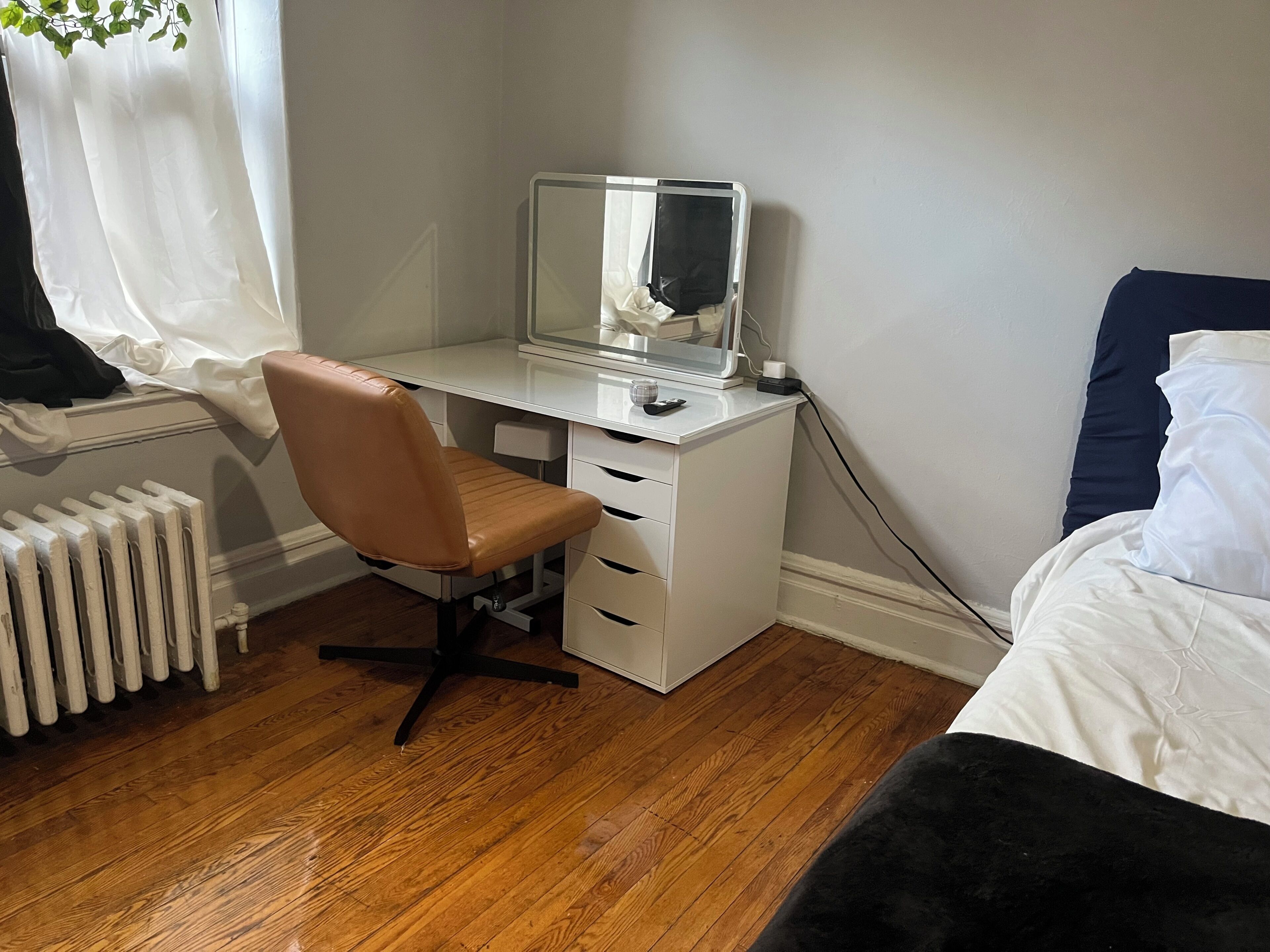 A bedroom with a bed, a desk with a computer, a chair, a radiator, and a window with curtains.
