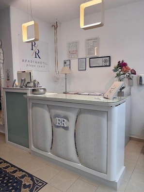 A reception desk with a logo, framed certificates, a lamp, and a vase with flowers. - Apartahotel Playa Conil (Conil de la Frontera)