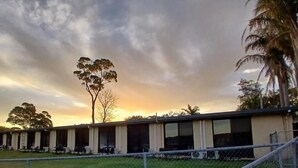 Uma fileira de edifícios térreos com grandes janelas, possivelmente um hotel ou hostel, com uma área gramada na frente e árvores ao fundo.