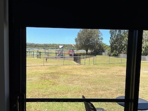 A view through a window of a grassy outdoor area with a playground and a fenced sports field. - Waterview Motel Sussex Inlet (Sussex Inlet)