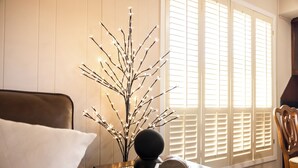 Una habitación con una silla de madera, una mesa con una rama decorativa y una ventana con persianas.