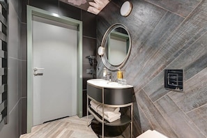 Une salle de bain moderne avec un mur recouvert de carreaux gris, un miroir rond, un meuble-lavabo noir et une porte blanche.