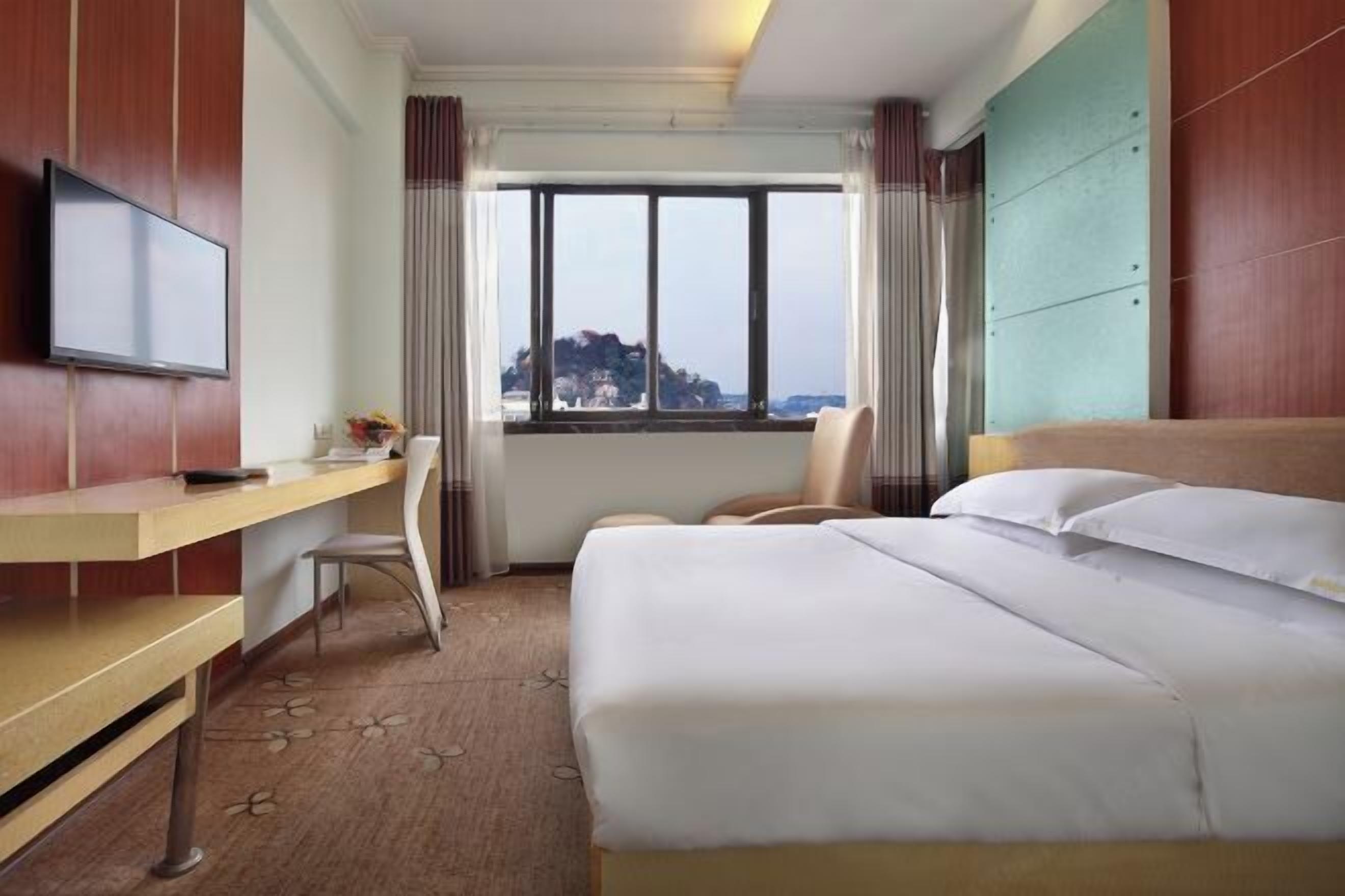 A hotel room with a bed, a desk, a chair, a TV, and a window with a view.