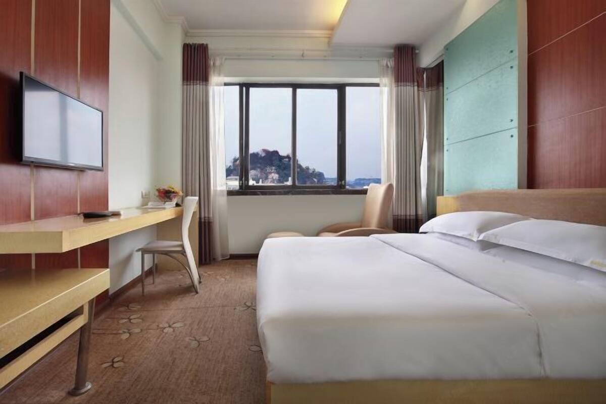 A hotel room with a bed, a desk, a chair, a TV, and a window with a view.